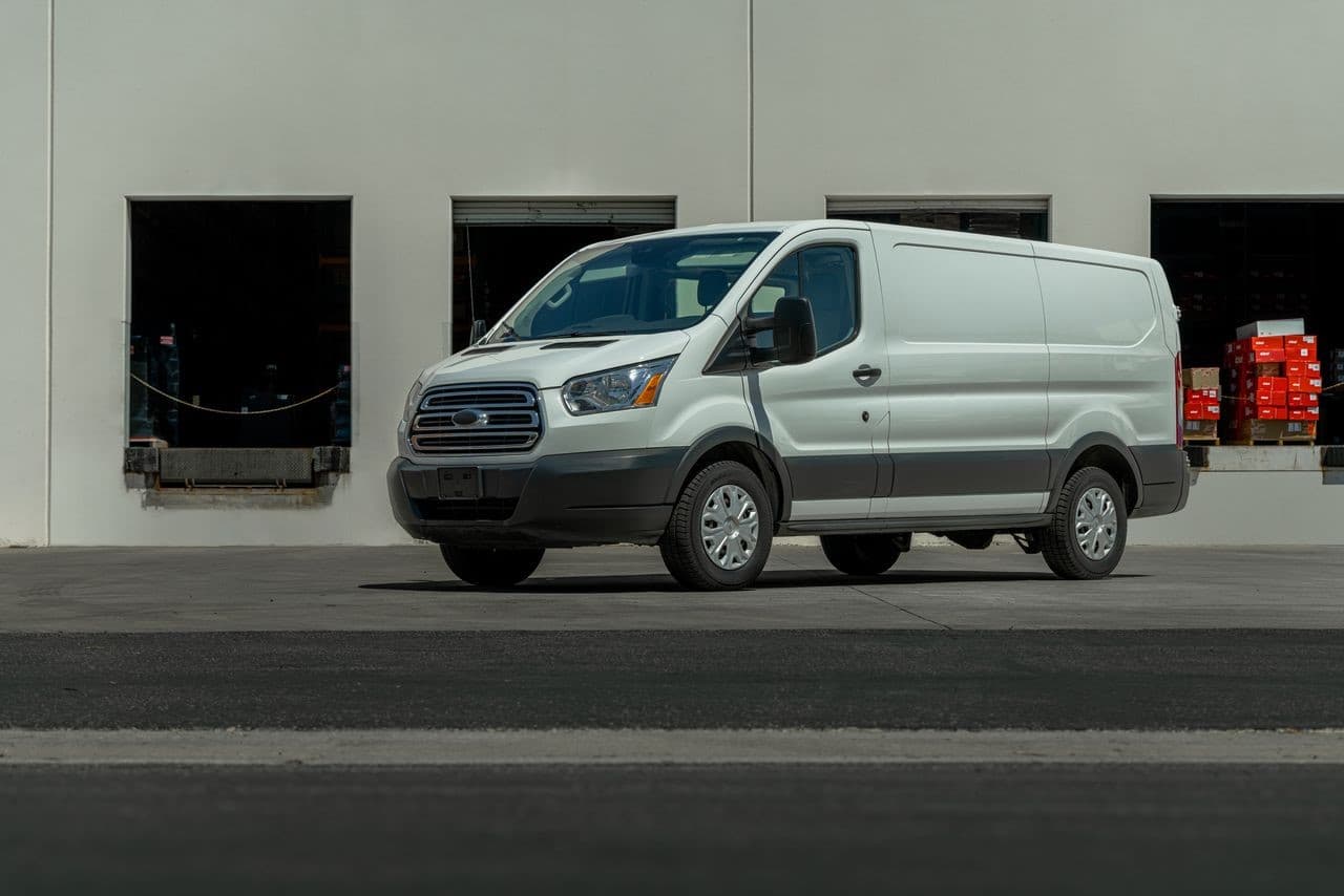 Furgoneta Ford Transit blanca estacionada frente a un edificio industrial con muelles de carga y caj