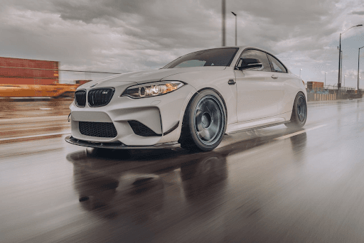 BMW M2 blanco manejando sobre pavimento mojado con fondo urbano y nubes grises, capturado en movimie
